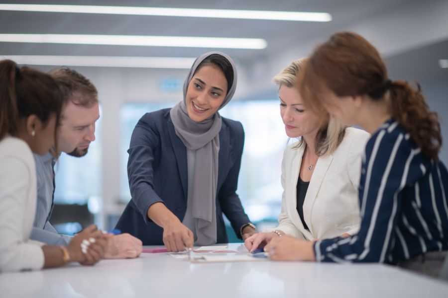 A woman wearing a hijab leads a discussion with a group of multi-ethnic businesspeople.