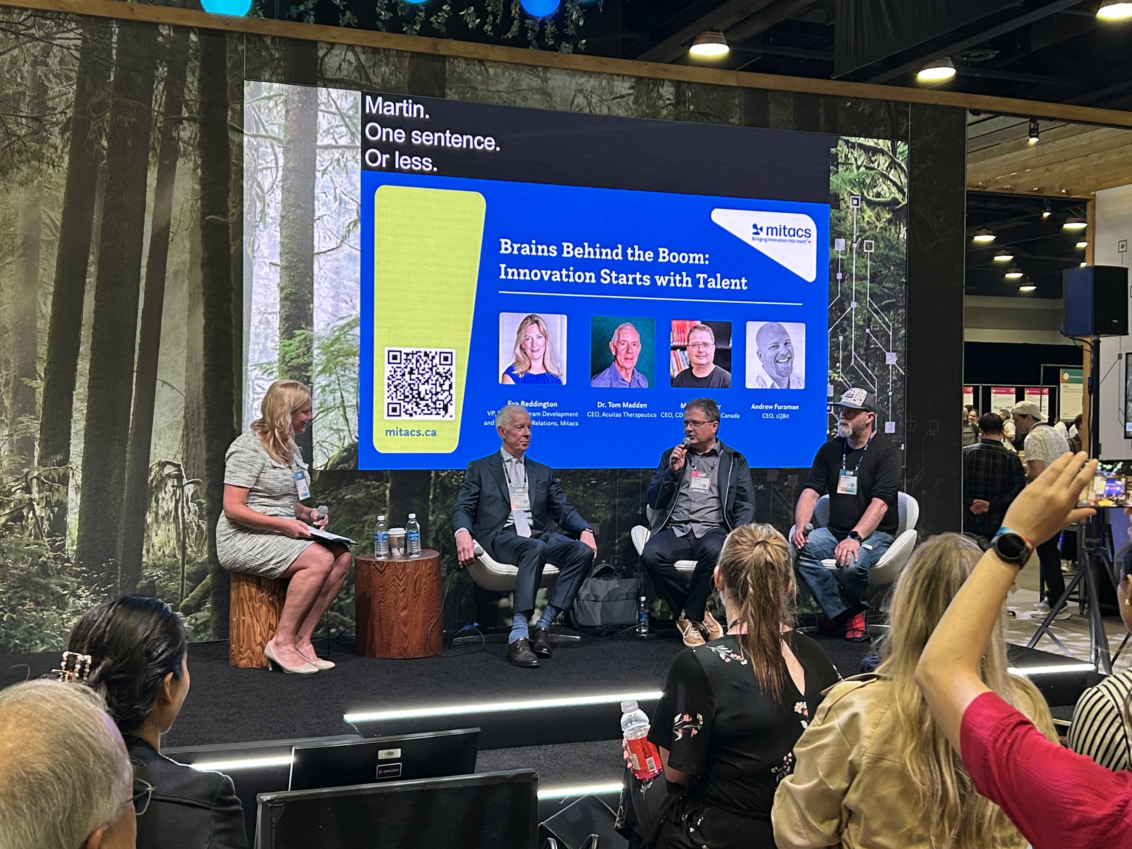 Five people sit on a stage in front of a large screen displaying Brains Behind the Boom: Innovation Starts with Talent, discussing how business R&D solutions can solve business challenges at an indoor event with a forest-themed backdrop.