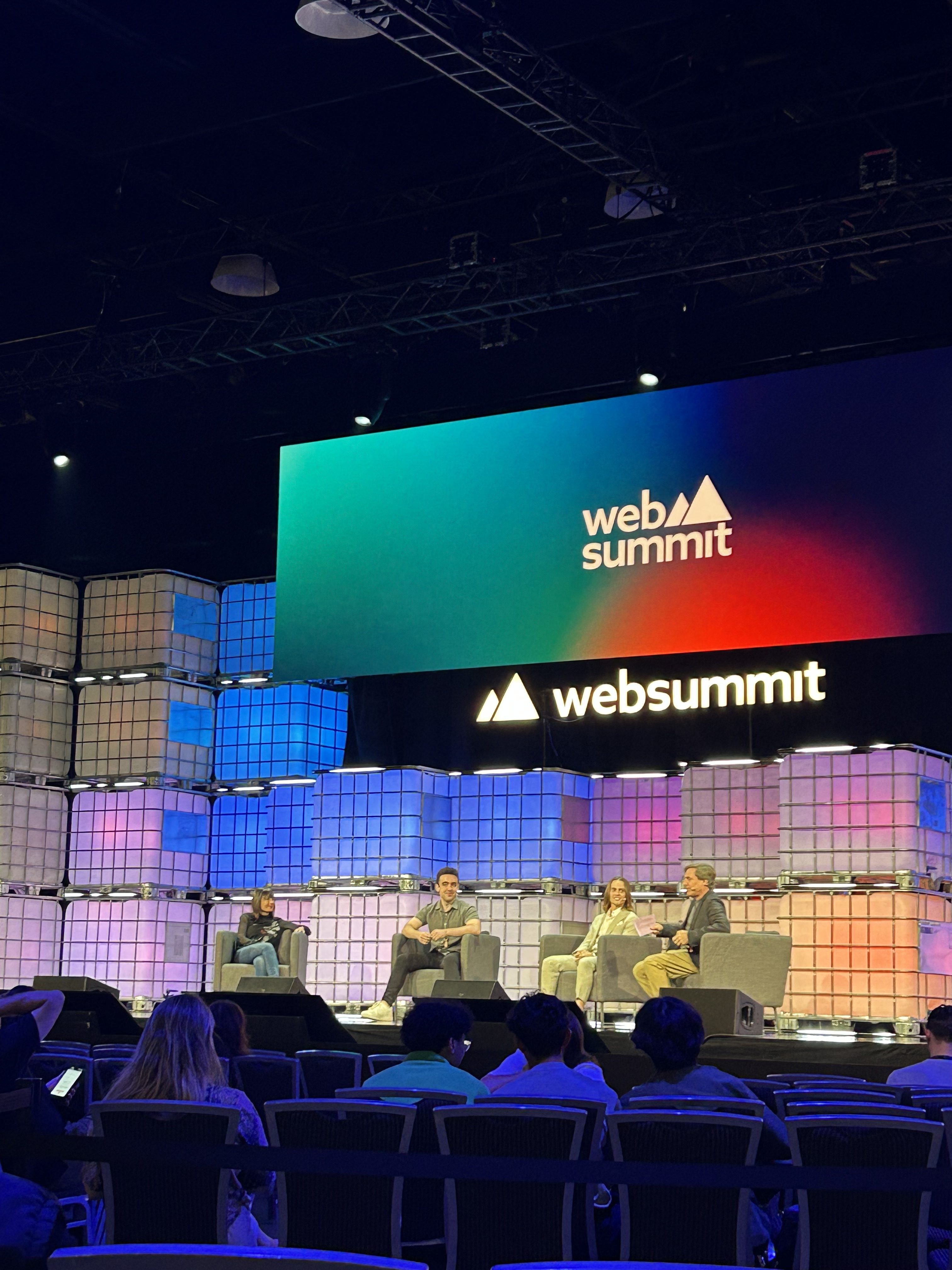 Four people sit on a stage in front of a large screen displaying Web Summit at a conference. The audience watches as they discuss business R&D solutions during a panel. The stage is decorated with stacked cube lights.