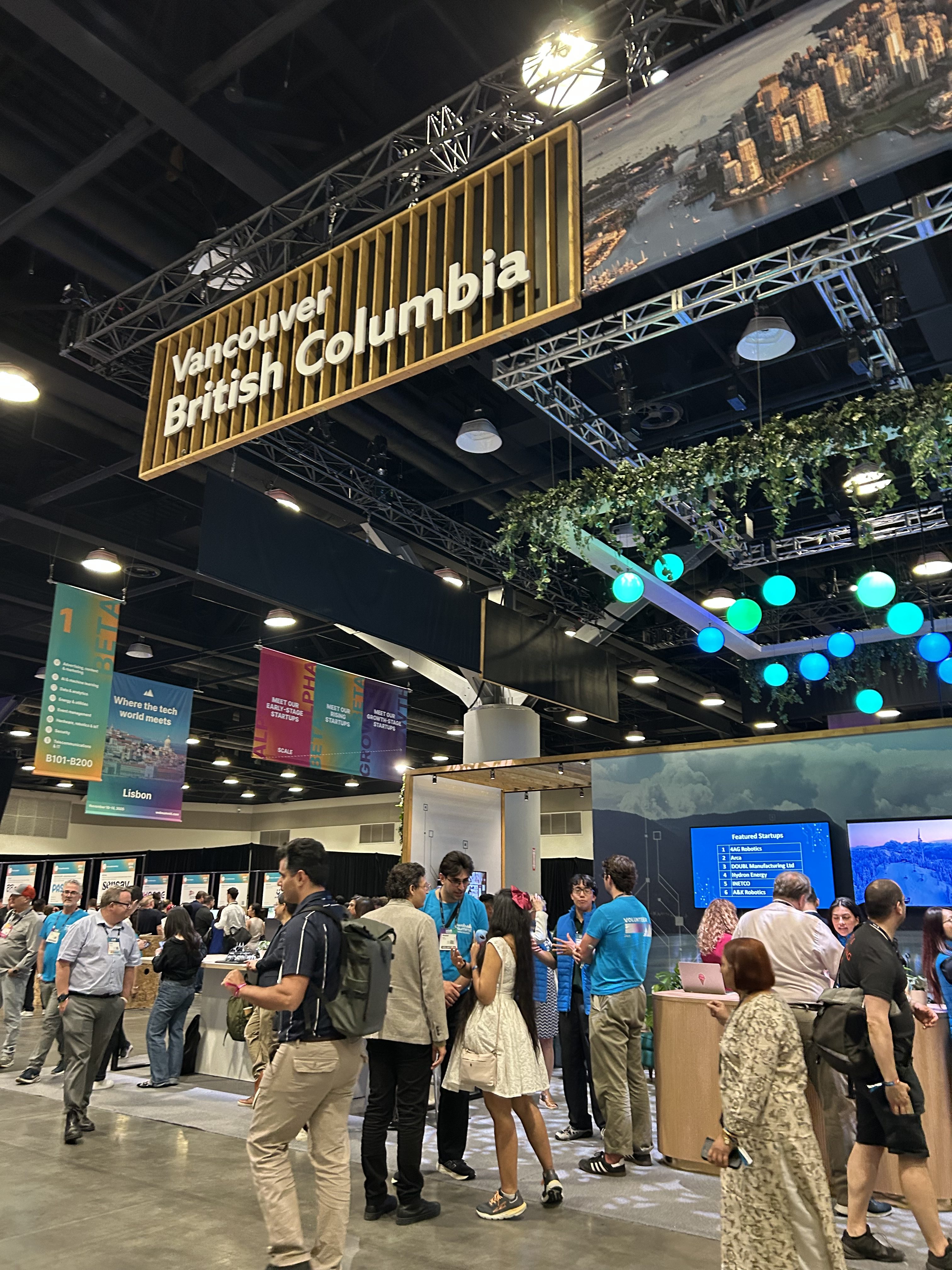 A busy convention center with people mingling near a booth featuring a large sign reading “Vancouver, British Columbia,” where experts discuss applied research Canada and innovative business R&D solutions amid hanging greenery and colorful lights overhead.