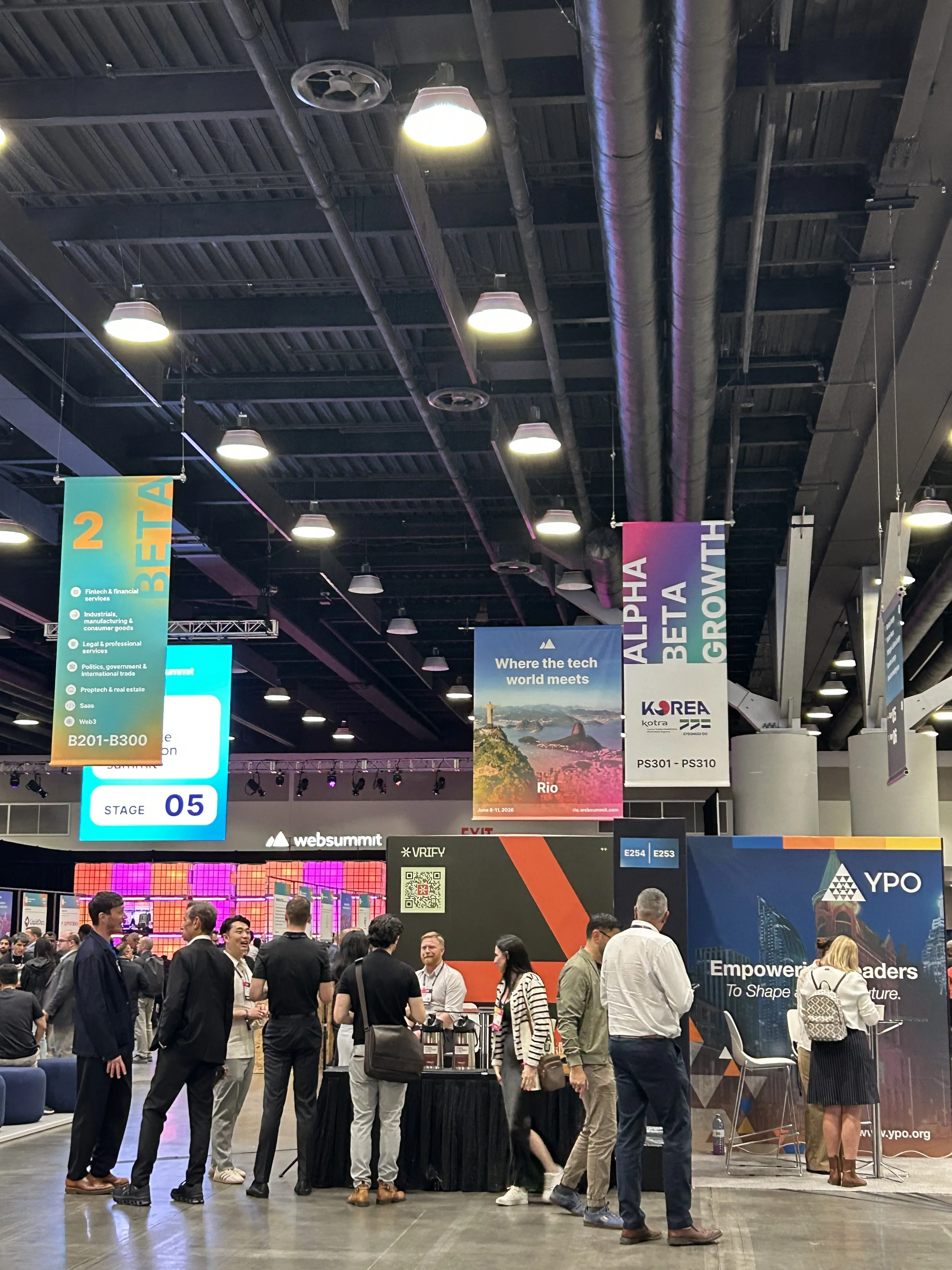 People gather and interact at a tech conference in a spacious hall. Colorful banners hang from the ceiling with words like ALPHA, BETA, and GROWTH, while booths highlight applied research Canada and business R&D solutions on digital screens.