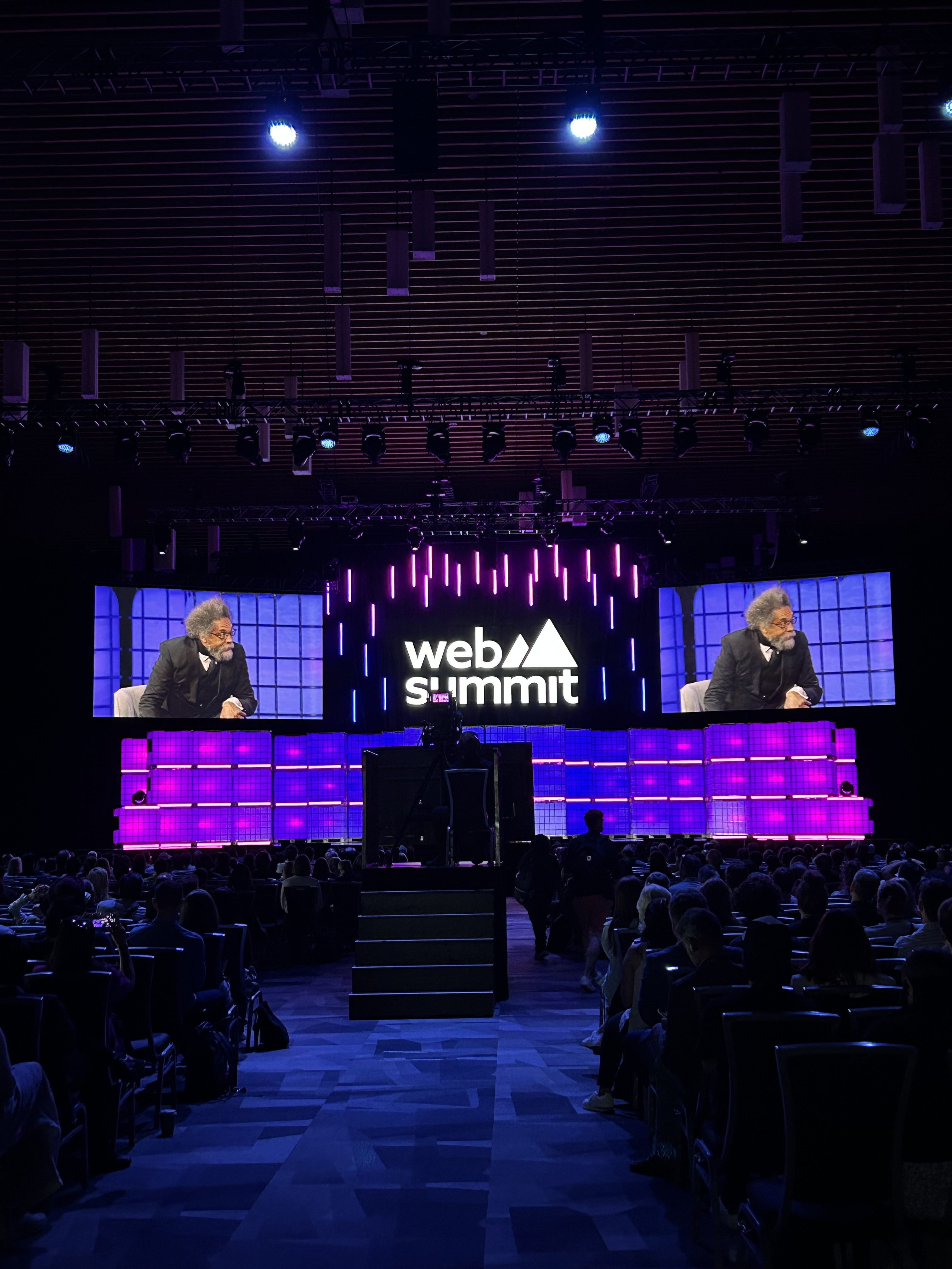 A speaker sits on stage at the Web Summit event, with large screens behind him displaying his image. The venue is dimly lit with purple lighting, as he discusses co-funded R&D projects and business R&D solutions before an attentive audience.