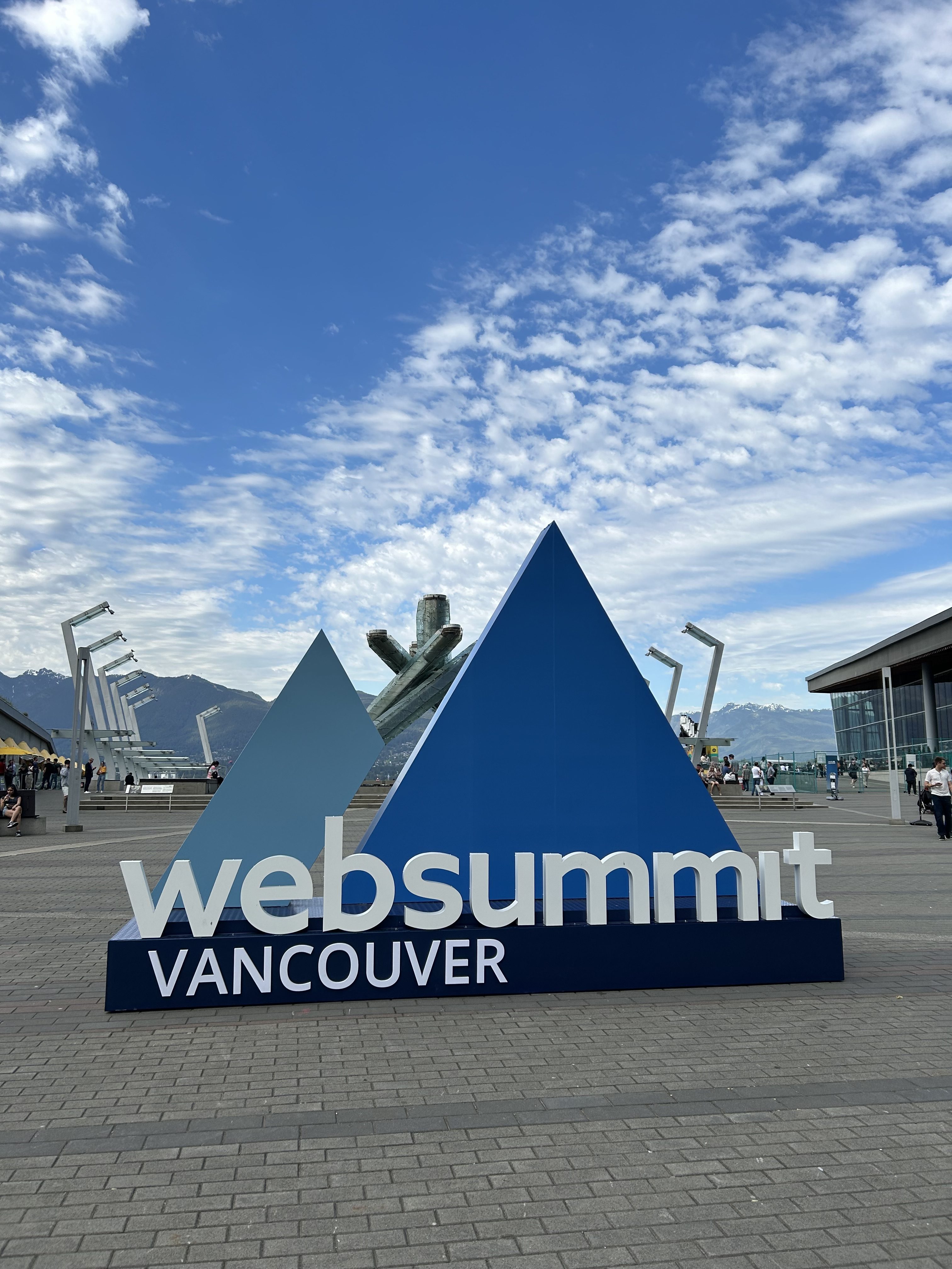 Large blue geometric sculpture with text websummit VANCOUVER stands outdoors on a plaza, framed by mountains and a modern building. Nearby, people walk—underscoring how Mitacs innovation funding can help solve business challenges in vibrant settings like this.