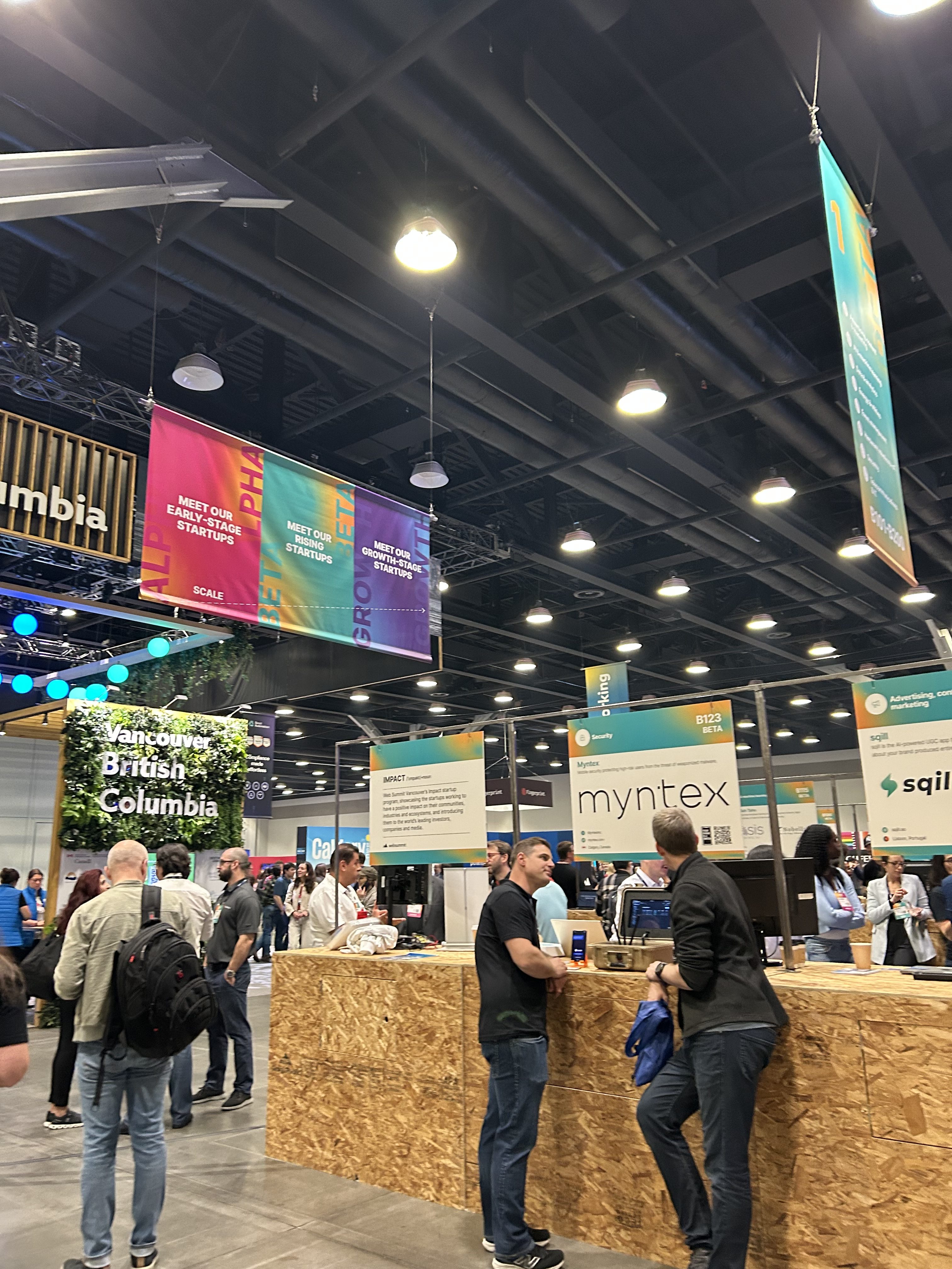 People gather at an indoor convention with high ceilings and bright lights. Various booths, including one for “myntex,” showcase business R&D solutions, with colorful banners and signs for “British Columbia” hanging above.