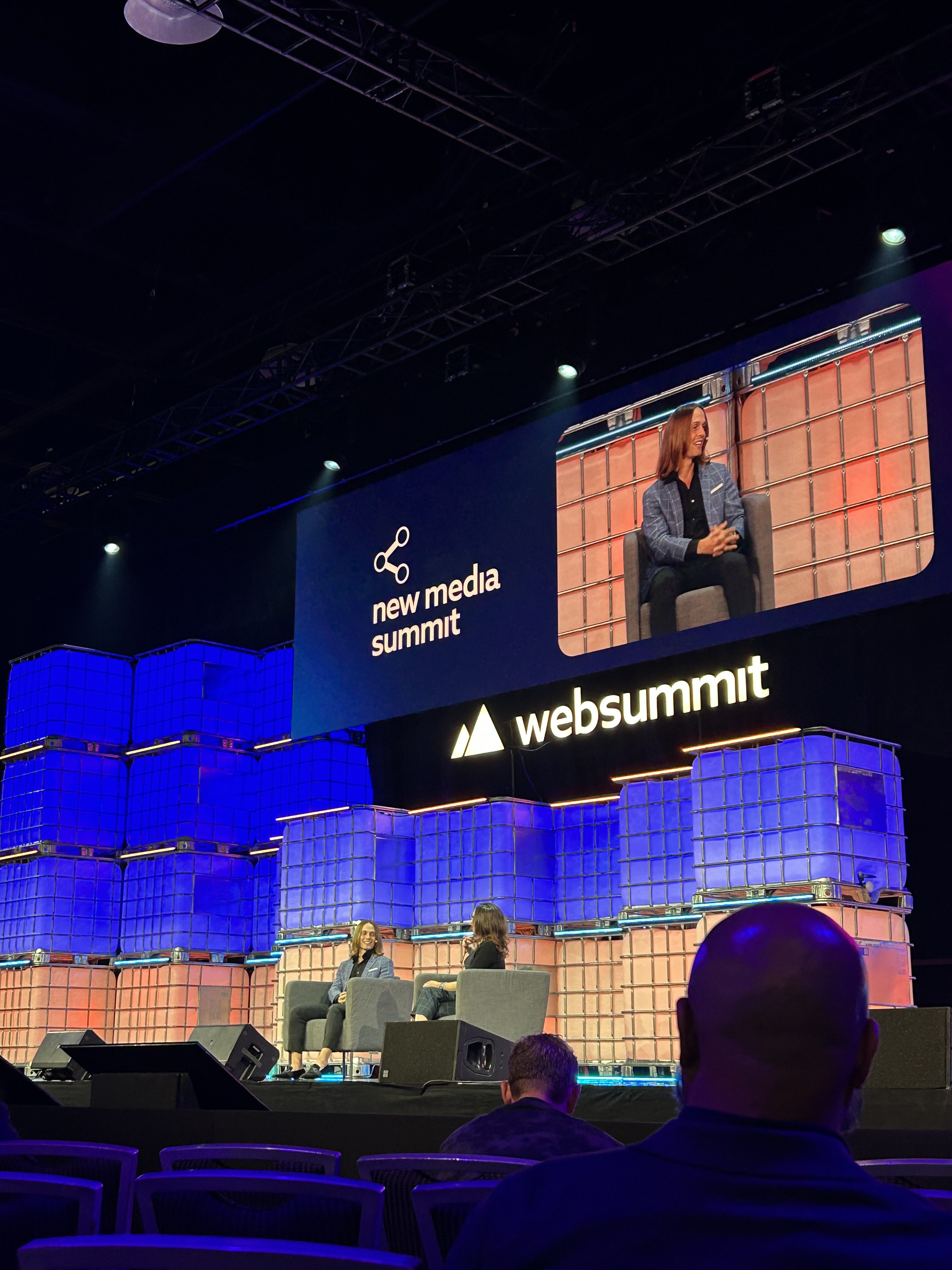 A stage at Web Summit features two people seated and talking, with one shown on a large screen above. The backdrop displays new media summit and websummit logos, highlighting how business R&D solutions can help solve business challenges. Audience members watch from the foreground.