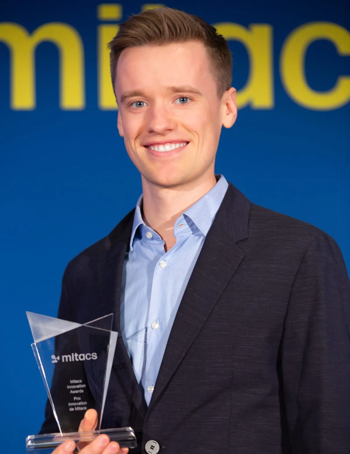 A smiling person in a navy blazer and light blue shirt holds a clear Mitacs award trophy, recognizing Canadian research achievements. The blue background features large partial yellow text, highlighting the prestige of Canadian research awards.
