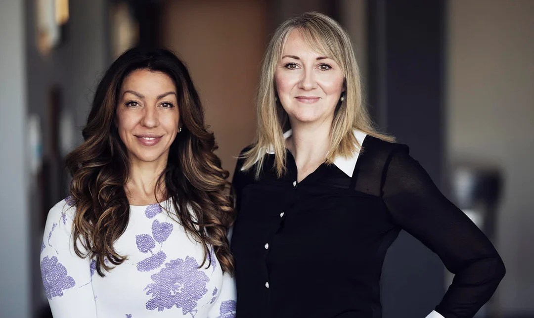Two women standing side by side indoors, smiling at the camera. One wears a white dress with purple floral patterns, and the other wears a black blouse with white cuffs and collar. The background is softly blurred.