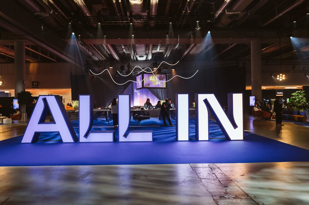 Large illuminated letters spelling ALL IN are displayed on a blue carpet in an indoor event space, with people and stage lights visible in the background.