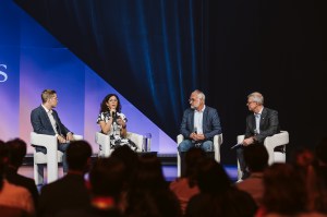 Four people sit on stage in armchairs, engaged in a panel discussion on innovation. One woman speaks into a microphone while the three men listen. An audience watches from the foreground as blue and purple lighting sets the scene.