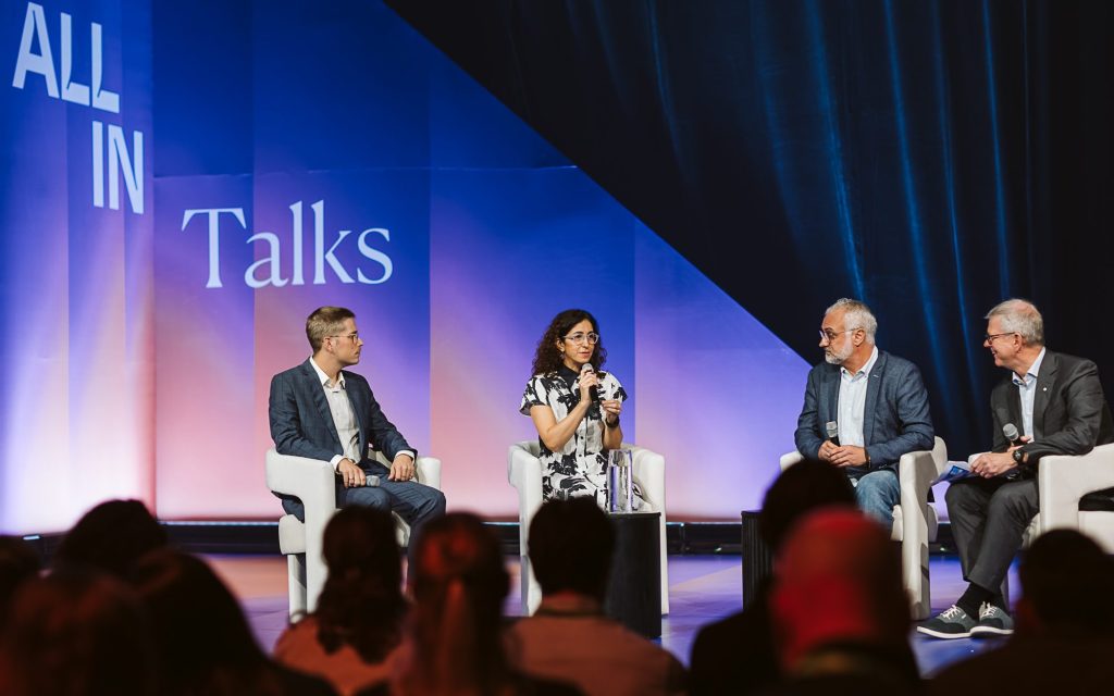 Four people sit on stage in armchairs, engaged in a panel discussion. The background features the words ALL IN Talks in large letters. Audience members are visible in the foreground, watching the conversation.