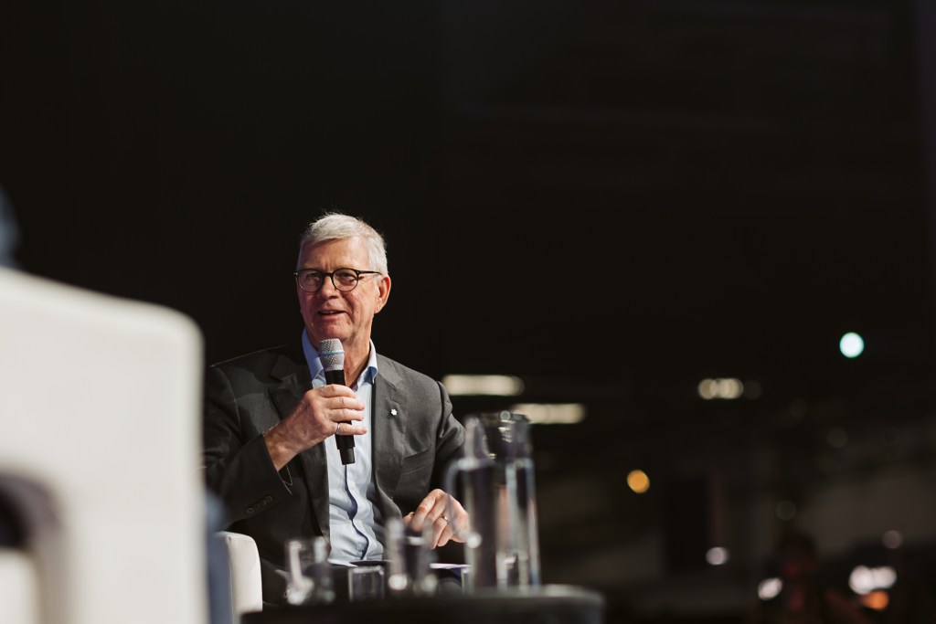 An older man in a suit and glasses speaks into a microphone while seated on stage. A glass water bottle and glasses are on the table in front of him, and the background is dark and out of focus.