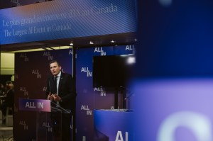 A man in a suit speaks at a podium labeled ALL IN at a tech event, with bilingual signage above reading The Largest AI Event in Canada, where entrepreneurs and an innovation network gather to solve business challenges.