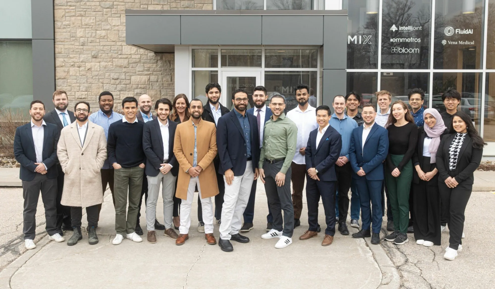 A diverse group of about 25 professionally dressed people stand smiling in front of a modern building with large windows and company logos, posing for an outdoor group photo on a cloudy day.