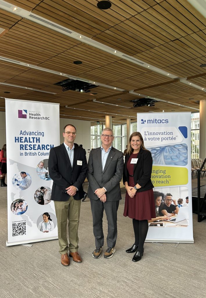 Trois personnes en tenue d’affaires se tiennent et sourient à l’intérieur entre deux bannières pour Health Research BC et Mitacs lors d’un événement professionnel, avec des sièges vides et de grandes fenêtres en arrière-plan.