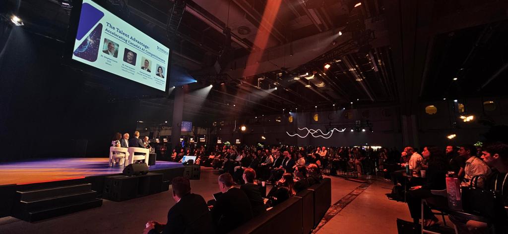 A large audience watches a panel of speakers seated on stage at a conference. A screen above displays event and speaker information. The venue is dimly lit, with spotlights highlighting the stage.