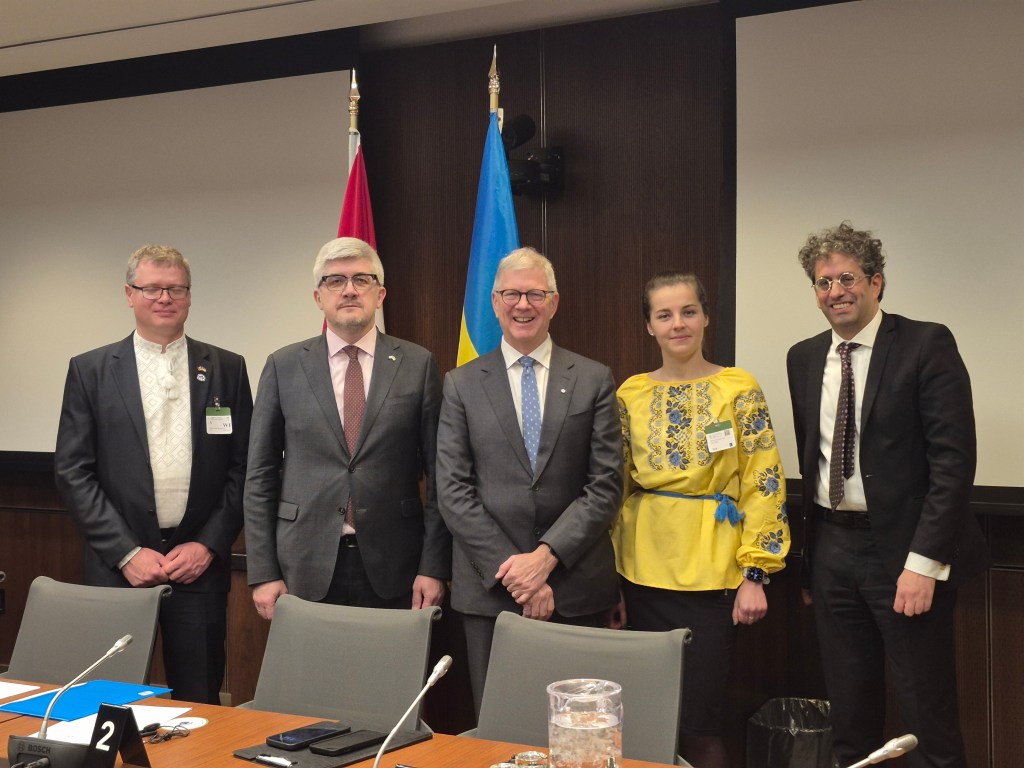Five people stand in a conference room, dressed in business and traditional attire, with Canadian and Ukrainian flags behind them. They smile for a group photo by the meeting table, highlighting the Mitacs Ukraine partnership.