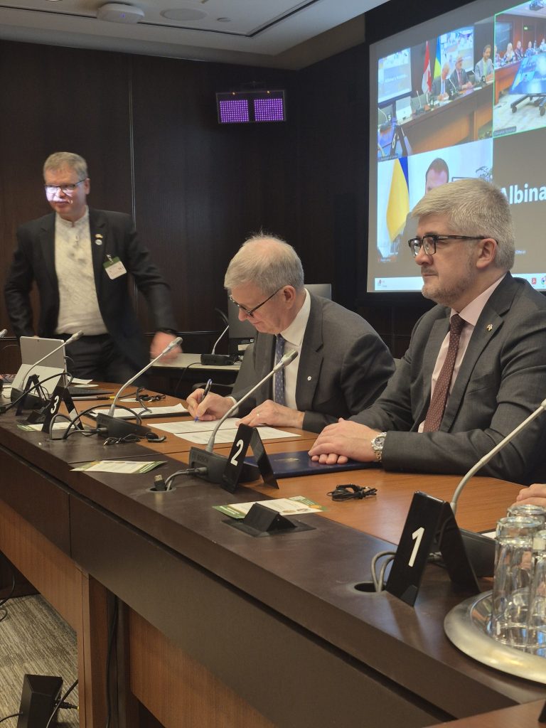 Three men in business attire sit at a conference table in a meeting room for the Mitacs Ukraine partnership; one signs a document while another sits beside him. Screens in the background display participants joining remotely.