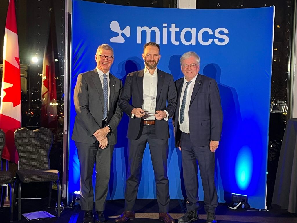 Three men in suits stand smiling in front of a blue Mitacs backdrop. The man in the middle, one of the 2025 Mitacs Innovation Awards Winners, holds a glass award. Two Canadian flags are visible on the left side of the image.