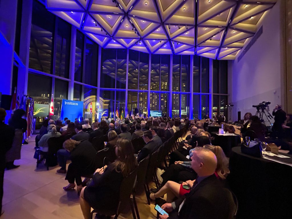 A large group of people sit facing a stage at an indoor evening event with floor-to-ceiling windows, geometric ceiling lights, and a cityscape visible outside as the 2025 Mitacs Innovation Awards Winners are celebrated under blue and yellow lighting.