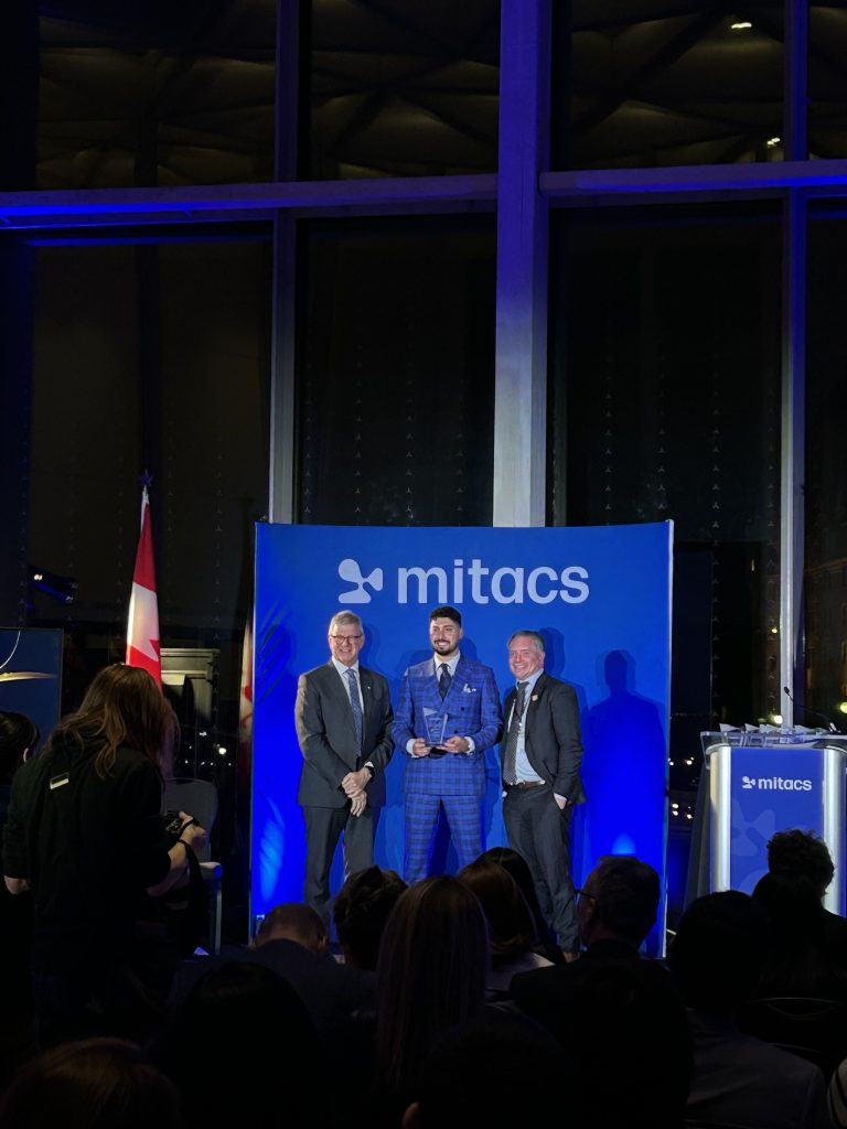 Three men in formal attire stand on a stage in front of a blue mitacs backdrop. One holds an award, celebrating as 2025 Mitacs Innovation Awards Winners. A Canadian flag is visible at the side, with an audience seated in the foreground.