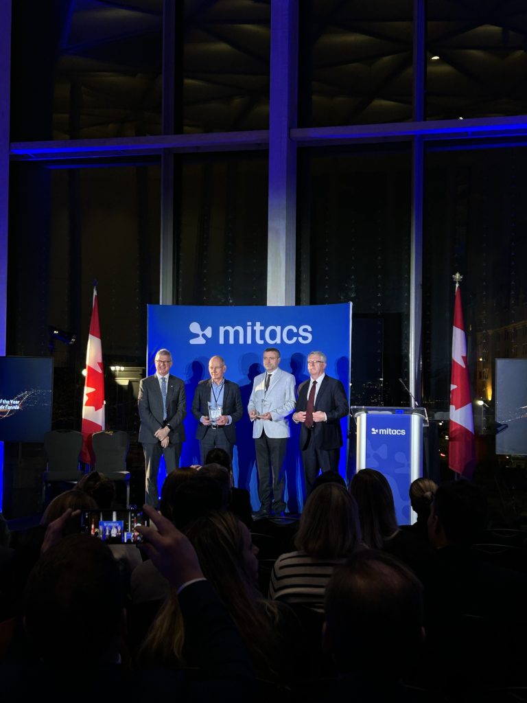 Four men in suits stand on a stage holding awards as the 2025 Mitacs Innovation Awards Winners, in front of a blue mitacs backdrop flanked by Canadian flags. The audience faces them, some taking photos, in a large venue with tall windows.
