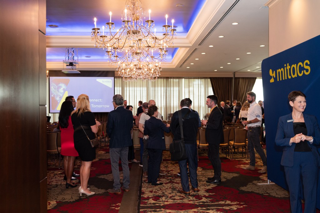 A group of people mingle in a conference room with chandeliers and a red-patterned carpet. A projector screen displays a presentation, and a blue sign with mitacs is visible on the right.