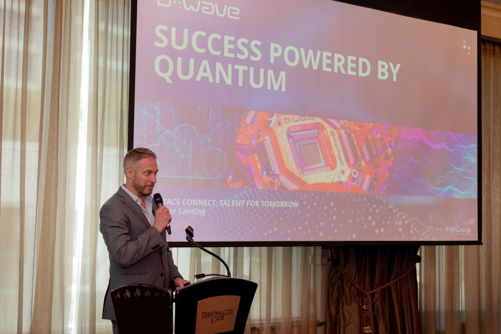 A man in a gray suit speaks at a podium in front of a large screen displaying the text SUCCESS POWERED BY QUANTUM at an event in a bright room with tall curtains.