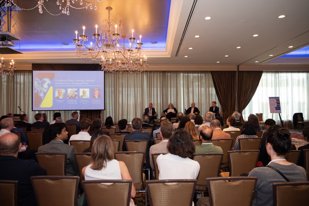 An audience watches a panel discussion in a conference room with chandeliers. A projection screen displays speaker photos and names at the front, where five people are seated on stage.