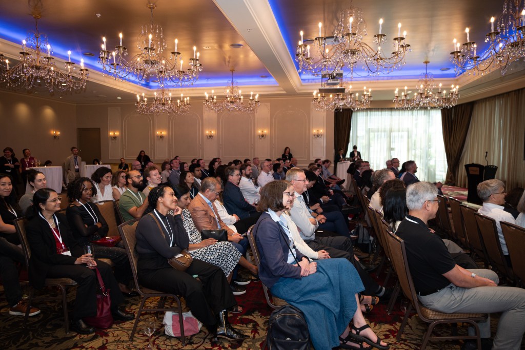 A diverse group of people sits attentively in rows of chairs at a conference in an elegant room with chandeliers and large windows, listening to a speaker out of frame.