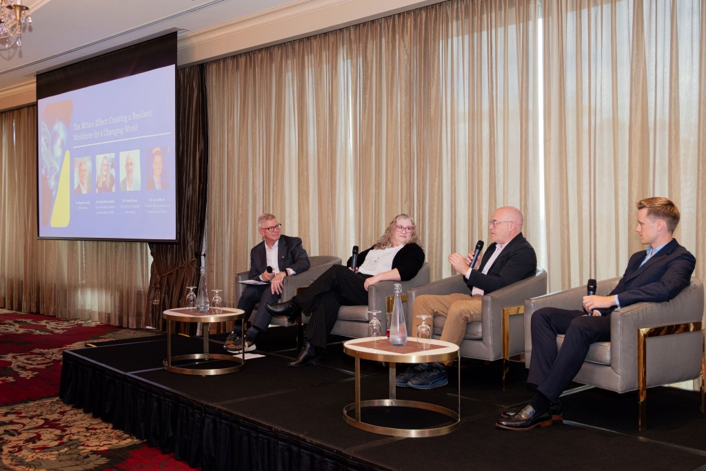 Four people sit on a stage having a panel discussion. Three men and one woman are seated in armchairs, with microphones, facing the audience. A presentation slide is displayed on a screen beside them.