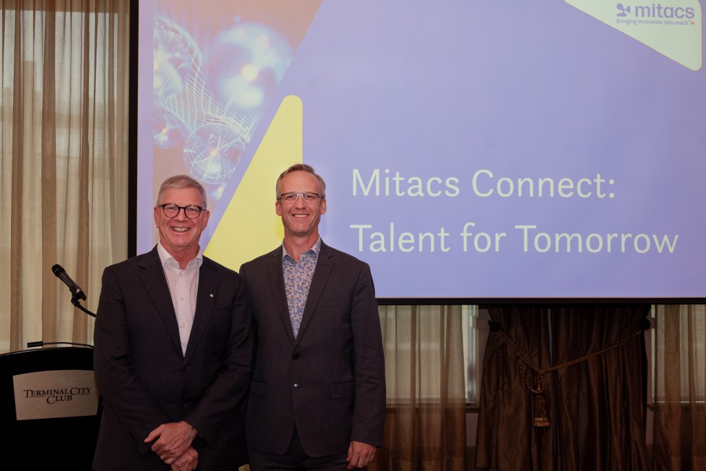 Two men in suits stand smiling in front of a screen displaying the words Mitacs Connect: Talent for Tomorrow at an event in the Terminal City Club. The background includes a Mitacs logo and abstract graphics.