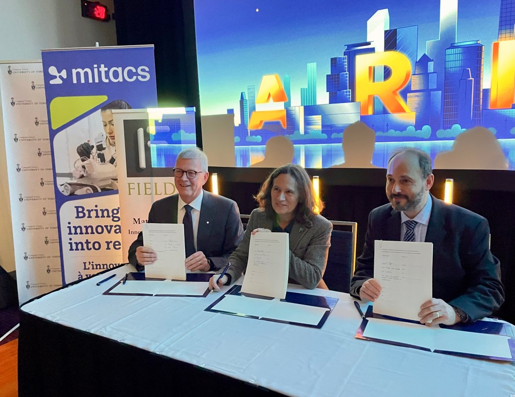 Three people sit at a table holding signed documents, posing for a photo at an event. Behind them are banners for Mitacs and Field, and a colorful screen displaying the word “AI.”.