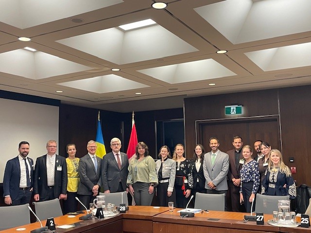 A group of sixteen people in business attire stand posing for a photo in a conference room, highlighting the Mitacs Ukraine partnership. The Ukrainian and Canadian flags hang behind them, with a wooden table set with microphones and nameplates.