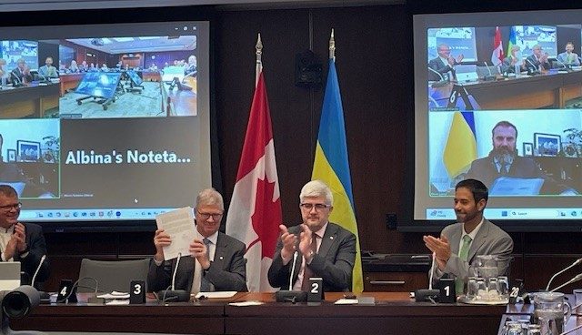Four men in suits sit at a conference table, clapping. Behind them are the Canadian and Ukrainian flags, highlighting the Mitacs Ukraine partnership. Video screens display a virtual meeting with participants and the text Albinas Noteta visible.