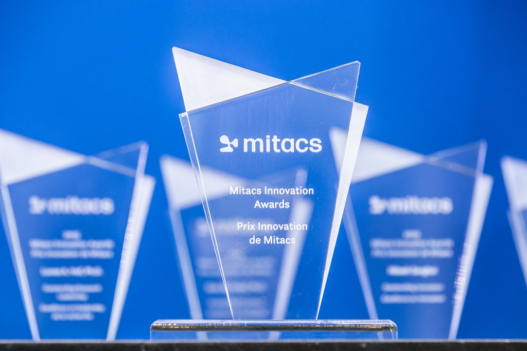 A clear glass trophy engraved with 2025 Mitacs Innovation Awards Winners stands in focus against a blue background, with similar awards blurred in the background.
