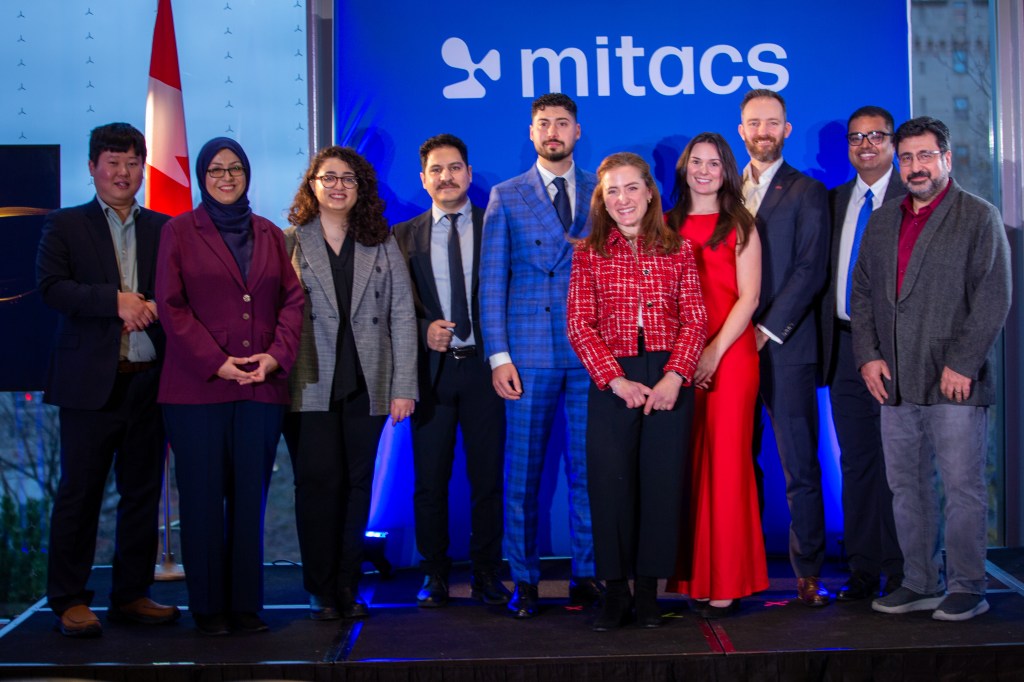 Ten people stand side by side, smiling at the camera in front of a blue Mitacs banner and Canadian flag. The group, dressed in business and formal attire, proudly represents the 2025 Mitacs Innovation Awards Winners.
