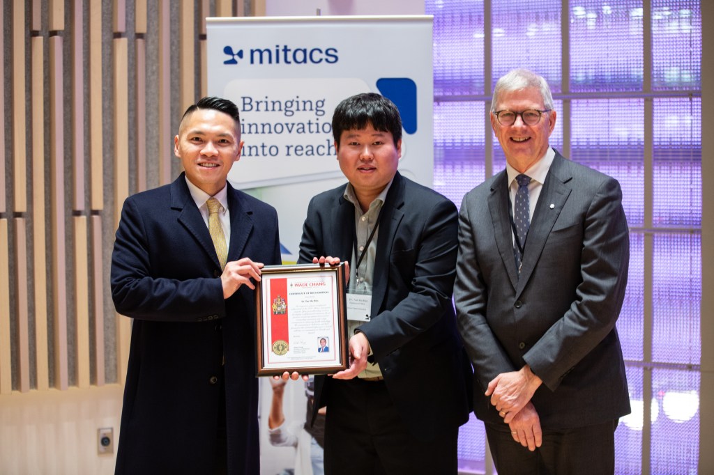 Three men in formal attire stand indoors; the man on the left holds a framed certificate with the man in the middle. A “mitacs” banner reading “Bringing innovation into reach” highlights them as 2025 Mitacs Innovation Awards Winners.