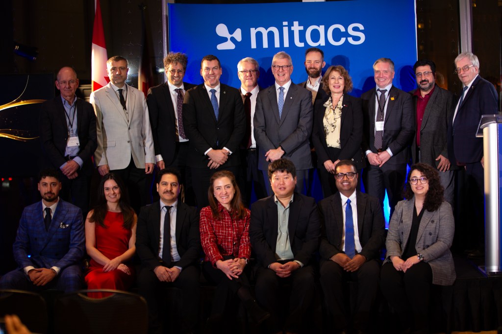 A group of eighteen people, including the 2025 Mitacs Innovation Awards Winners, pose for a photo in front of a blue Mitacs banner at an indoor event, with some seated in front and others standing behind them.