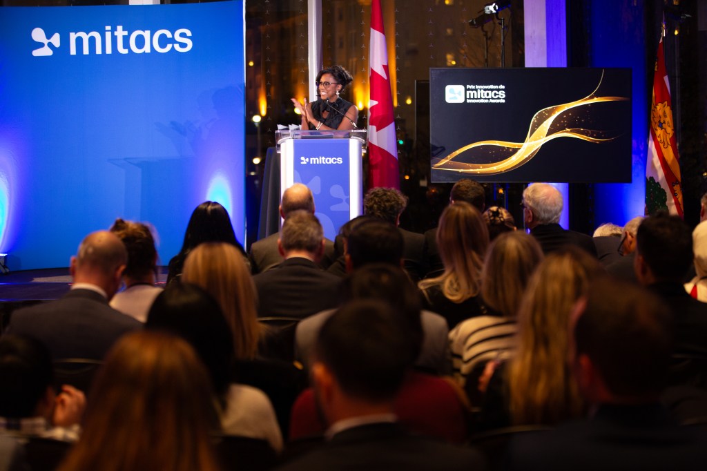 A speaker addresses an audience from a podium at a Mitacs event, with the 2025 Mitacs Innovation Awards Winners being celebrated. A Canadian flag stands nearby, and a large screen displays the Mitacs logo and a stylized graphic.