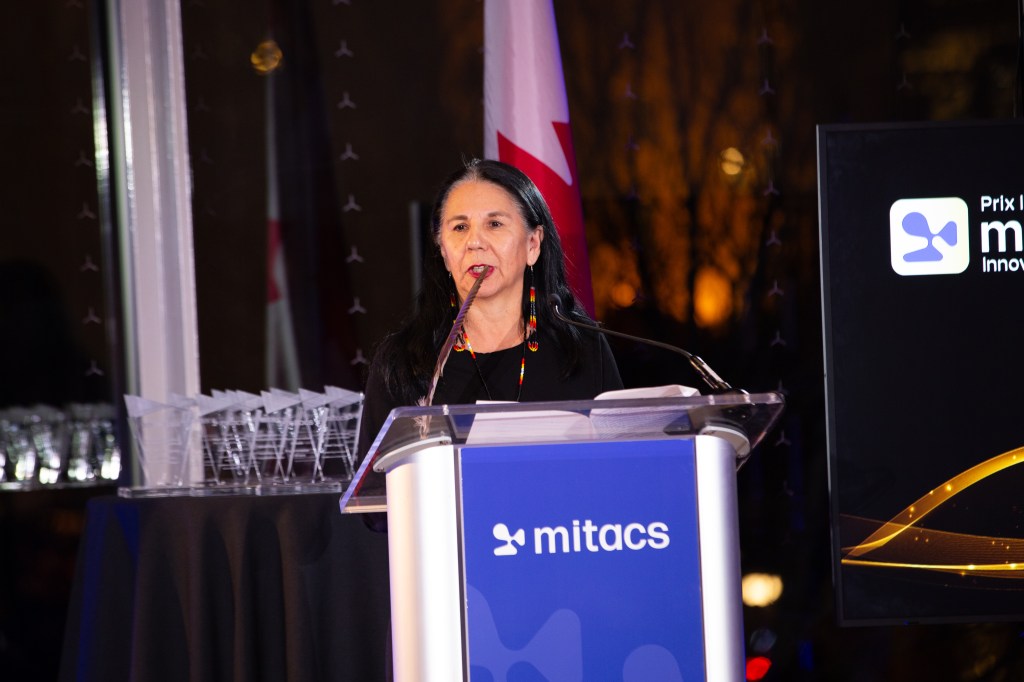 A woman, one of the 2025 Mitacs Innovation Awards Winners, stands at a podium with a microphone.