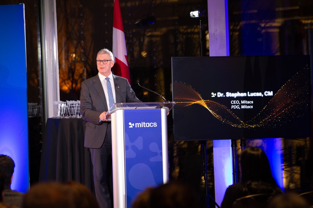 A man in a suit speaks at a podium with a Mitacs logo. Behind him, a screen displays Dr. Stephen Lucas, CM, CEO, Mitacs and a Canadian flag is visible—announcing the 2025 Mitacs Innovation Awards Winners to an attentive audience.
