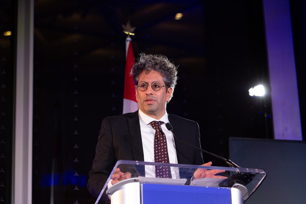A man with curly hair and glasses, wearing a suit and polka-dot tie, speaks at a podium with a microphone. A Canadian flag is visible in the background at what appears to be the 2025 Mitacs Innovation Awards Winners formal indoor event.