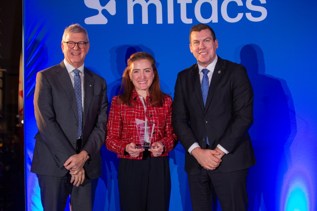 Three people in formal attire stand smiling in front of a blue background with the Mitacs logo. The woman in the center, one of the 2025 Mitacs Innovation Awards Winners, proudly holds a glass award.