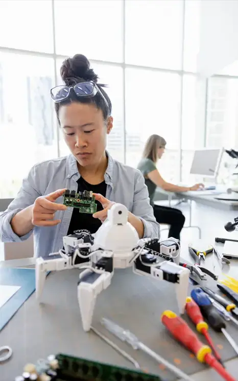 A woman examines a circuit board at a desk with a robotic device and tools, highlighting Canadian innovation leadership, while another person works at a computer in the background of a bright, modern workspace.