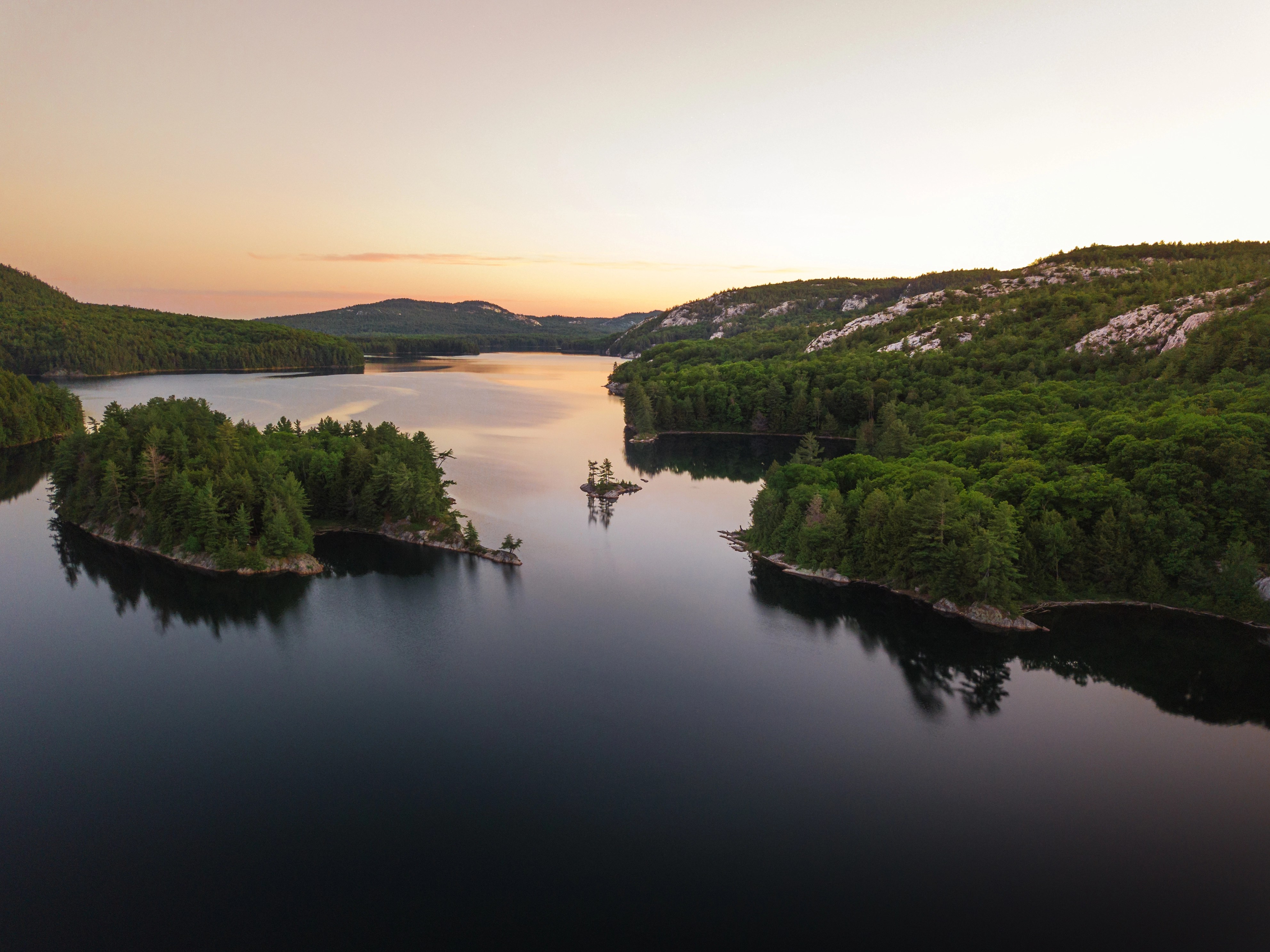 A tranquil lake surrounded by forested hills at sunset, with small tree-covered islands reflected in the calm water and a soft orange and pink sky—a scene echoing the harmony fostered by research partnerships and innovation funding Canada supports.
