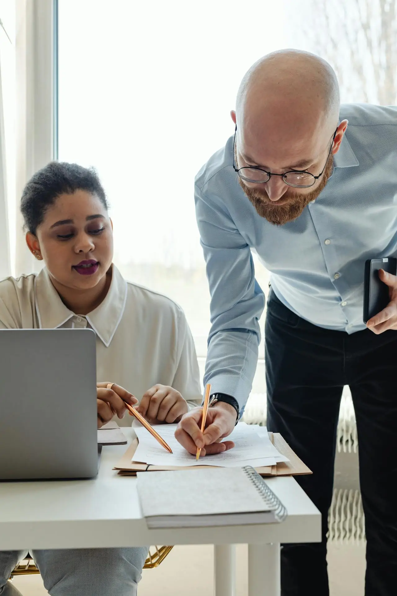 A woman at her desk with a laptop listens as a man beside her points to a document and holds a tablet. Both discuss innovation funding in a bright office, exploring ways to leverage their R&D budget for new opportunities.