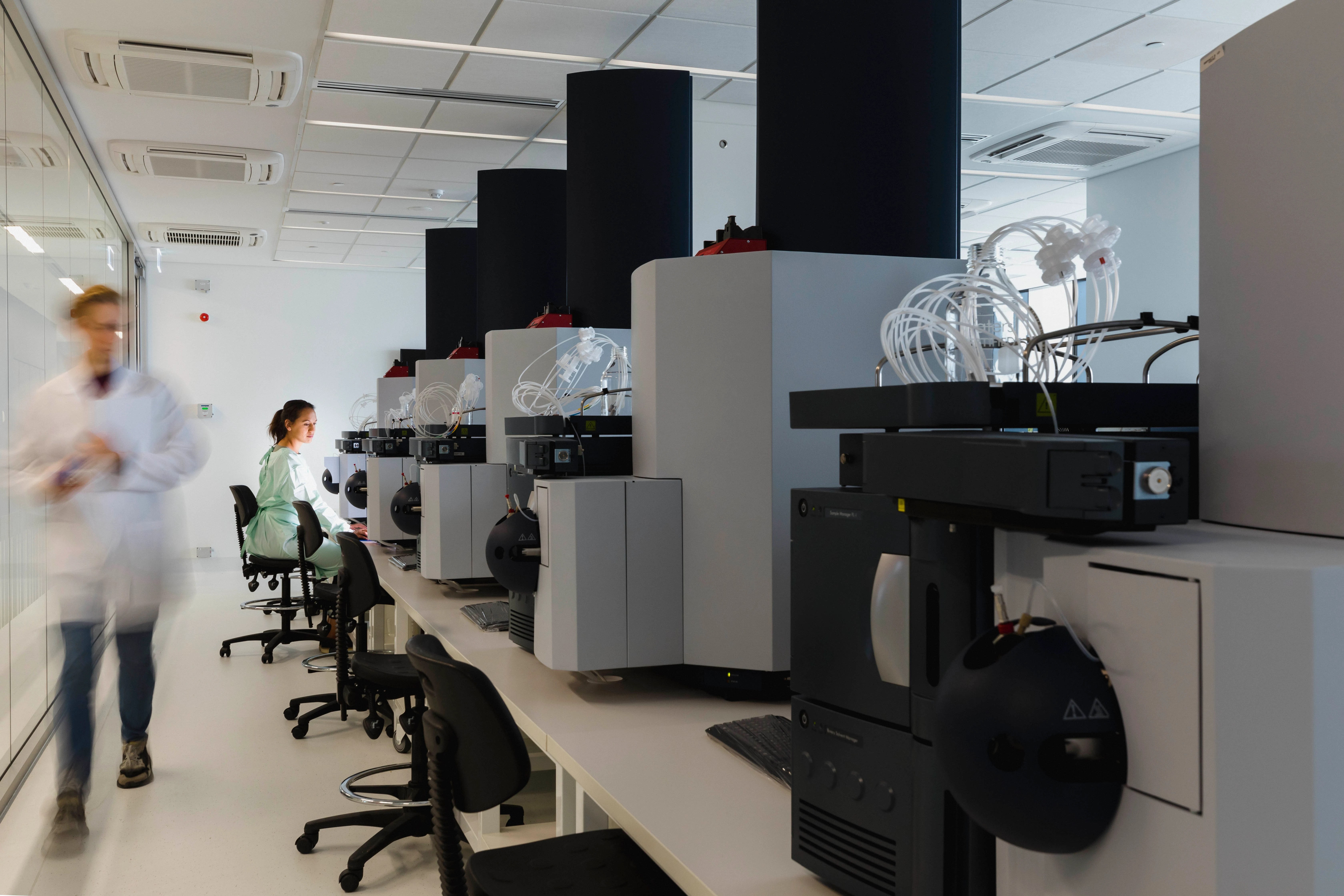 Scientists work at computers and machinery in a modern, brightly lit laboratory. One person sits at a desk, while another in a lab coat walks through the room, showcasing the talent development driving the Canadian innovation ecosystem.
