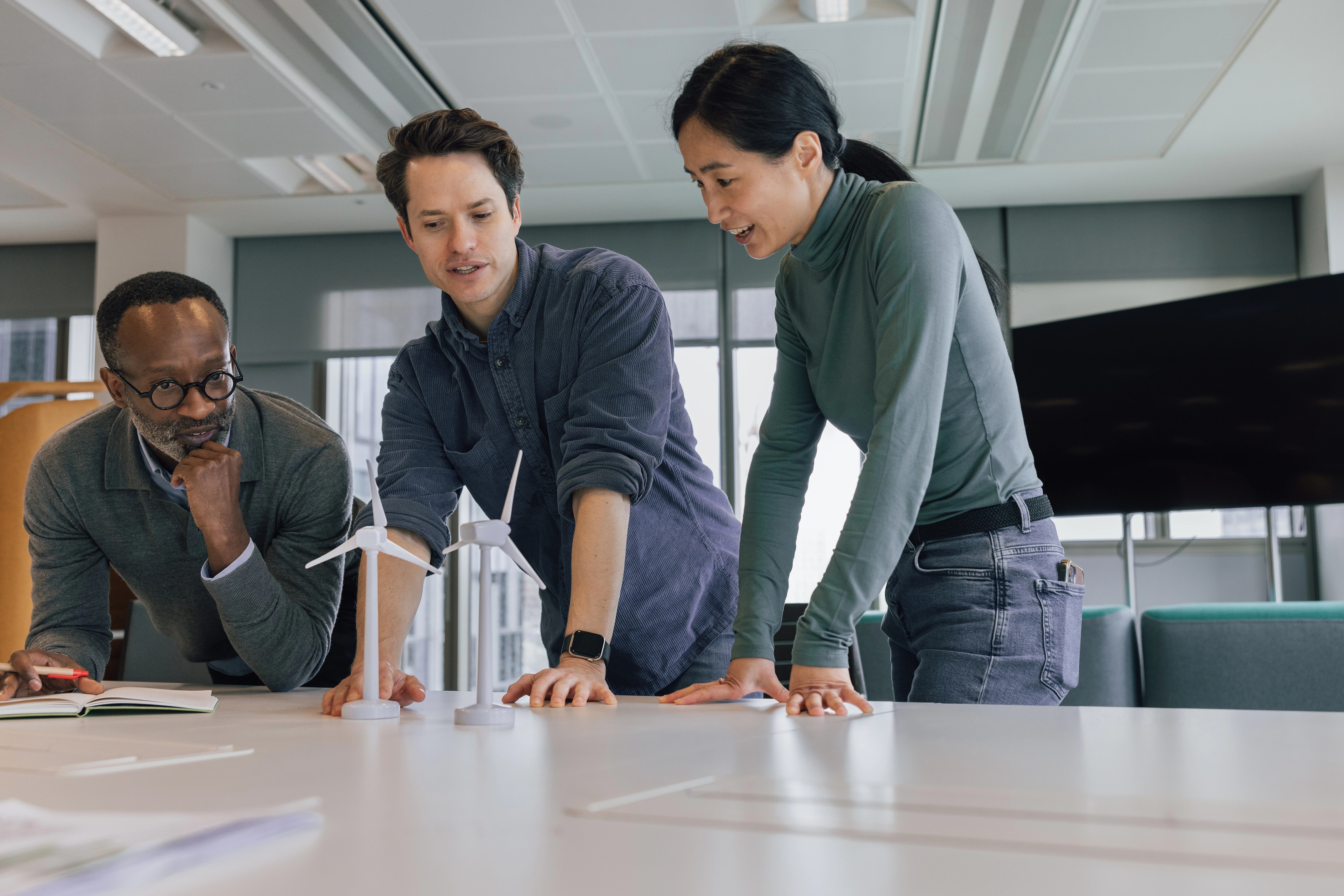 Three people stand around a table in an office, examining small wind turbine models and discussing ideas—demonstrating the spirit of Canadian innovation ecosystem and collaboration that drives renewable energy projects forward.
