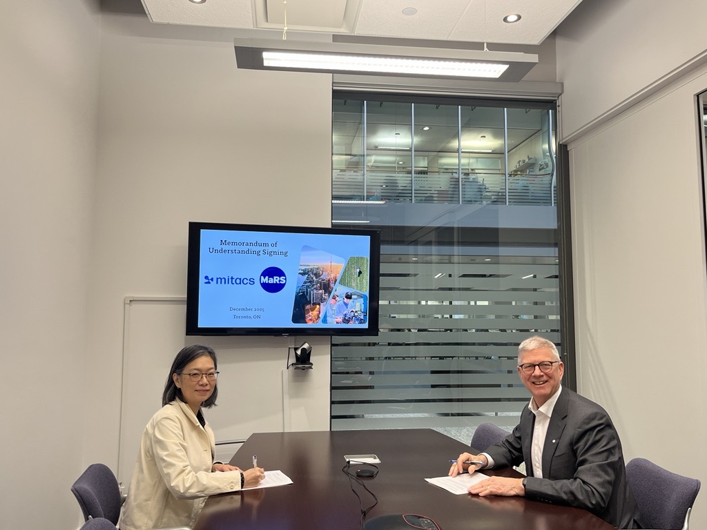 Two people sit at a conference table signing documents. A screen behind them displays “Memorandum of Understanding Signing” with Mitacs and MaRS logos. An office area with glass walls and people is visible in the background.