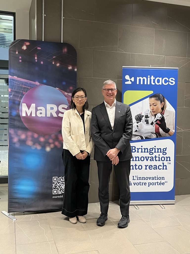 Two people stand smiling in front of banners for MaRS and Mitacs. The Mitacs banner features a scientist using lab equipment and the words Bringing innovation into reach. The MaRS banner has a QR code and a blurred light background.